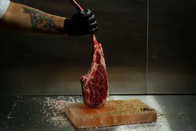 Persona con guante sosteniendo un corte de carne cruda sobre una tabla de preparación.